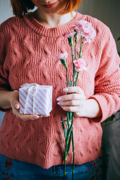 Frau im korallenroten Mädchen der wollenen Strickjacke, das Geschenkbox mit Band und frischen Grünblättern hält und rosa Blumen blüht