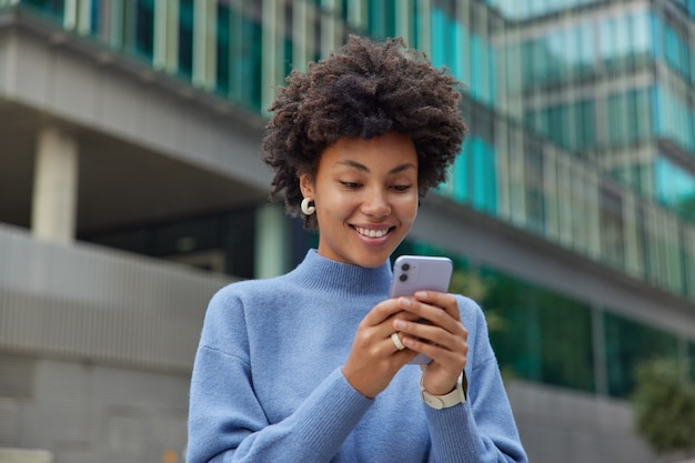 Frau im lässigen blauen Pullover benutzt das Handy, um online mit ihrem Freund zu chatten, der sich über die Benachrichtigung freut, und verbringt ihre Freizeit damit, in der Stadt zu bummeln