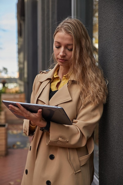 Frau im Mantel mit Tablet im Freien an einem sonnigen Tag beim Lesen von E-Books, die einen Film ansehen