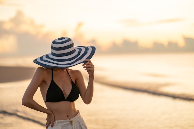 Frau im oberen Bikini und in der weißen langen Hose, die Hut am Strand mit einem schönen Sonnenaufgang trägt