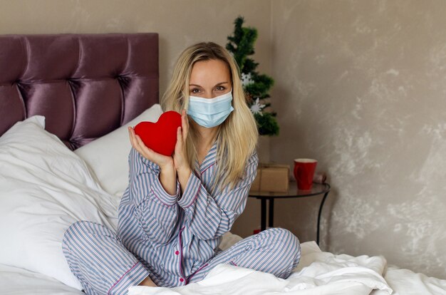 Frau im Pyjama in einer medizinischen Maske hält ein Herz, das Weihnachten auf dem Bett feiert