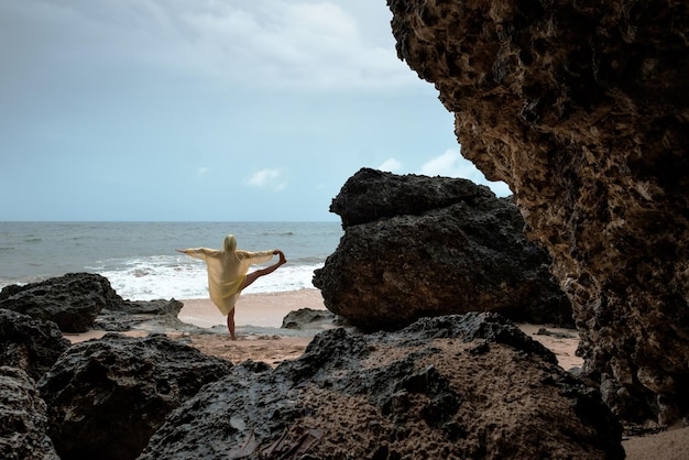 Frau im Regenmantel, die während des Sturms Balance-Yoga-Asanas am Meeresstrand macht