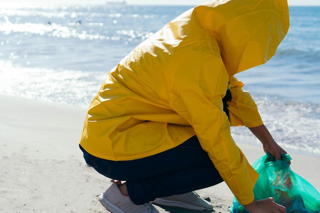 Frau im Regenmantel hebt Plastik gebückt am Strand auf