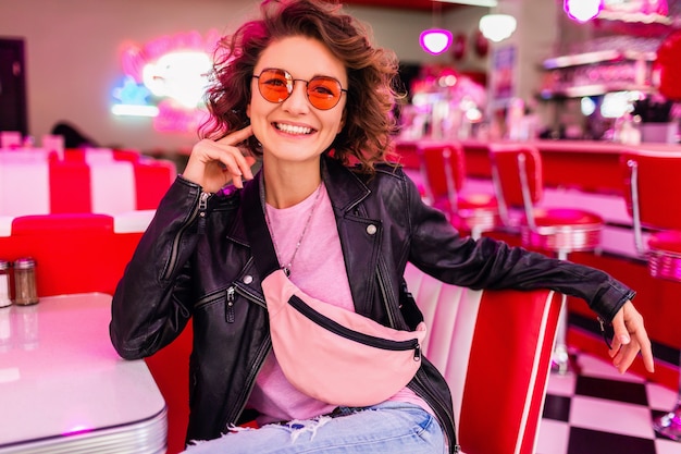 Frau im Retro-Vintage-Café der 50er Jahre am Tisch in schwarzer Lederjacke mit rosa Sonnenbrille und Geldbörse, die sich in fröhlicher Stimmung amüsiert