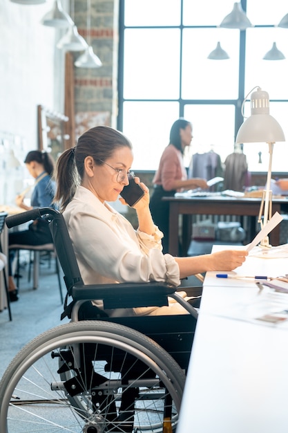 Frau im Rollstuhl, die durch Papier schaut und mit dem Kunden auf dem Smartphone spricht