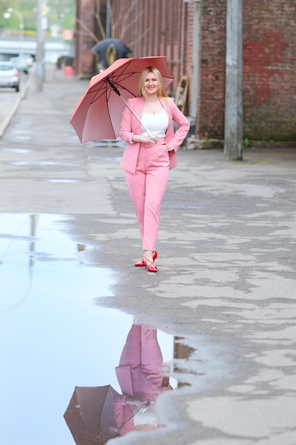 Frau im rosa Anzug mit Regenschirm gehend auf die Straße nach dem Regen