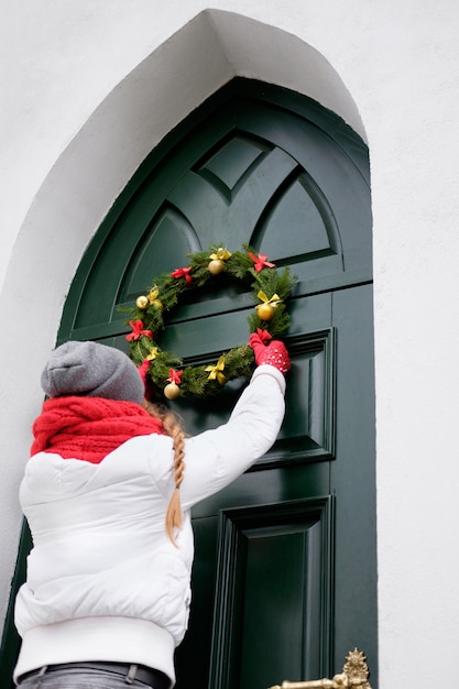 Frau im roten Schal und in der weißen Jacke, die Weihnachtskranz an der grünen Tür hängen