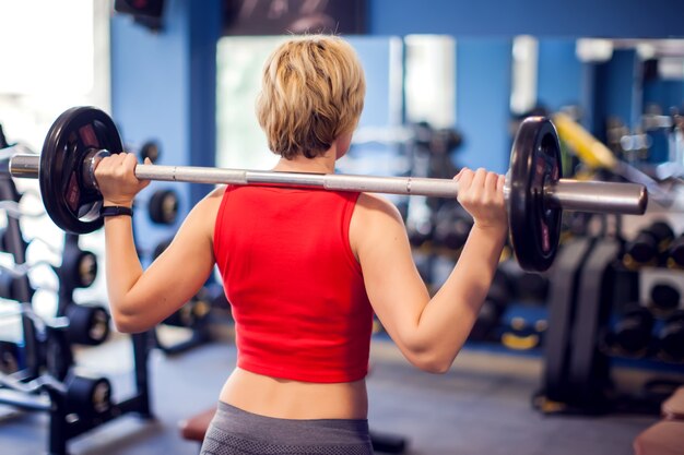 Frau im roten Top mit kurzen blonden Haaren, die sich darauf vorbereiten, Kniebeugen im Fitnessstudio zu machen. Menschen-, Fitness- und Lifestyle-Konzept