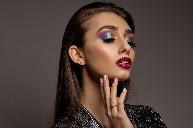 Frau im schwarzen glänzenden Kleid berührt ihr Gesicht und posiert auf grauem Hintergrund Luxus Make-up buntes Auge...