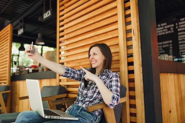 Frau im Sommercafé aus Holz im Freien, das mit Laptop-PC-Computer sitzt, Selfie-Aufnahme auf dem Handy macht und sich in der Freizeit entspannt Mobiles Büro