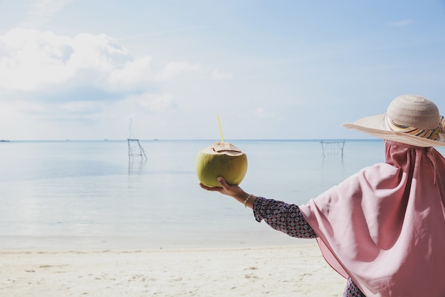 Frau im Sommerhut, die Kokosnuss am Strand mit schönem Naturhintergrund mit Kopienraum hält