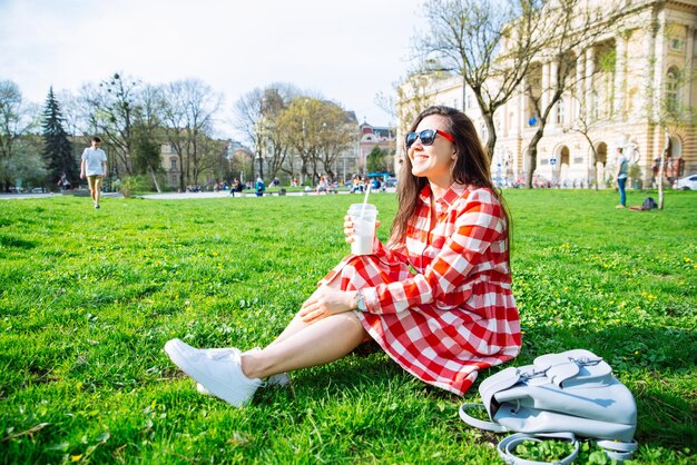 Frau im Stadtpark sitzt auf dem Boden und trinkt Smoothie