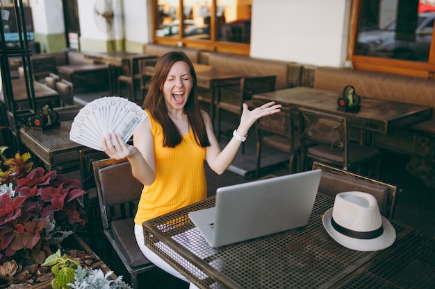 Frau im Straßencafé-Café im Freien, die mit einem modernen Laptop-PC-Computer sitzt, hält in der Hand einen Haufen Dollar-Banknoten