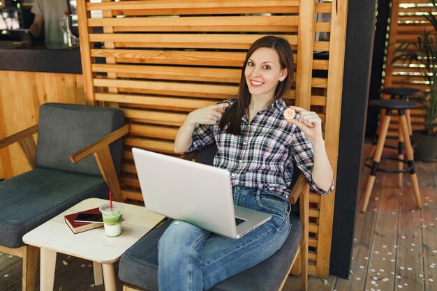 Frau im Straßencafé-Café im Freien, die mit Laptop-PC-Computer sitzt, Bitcoin halten, Metallmünze von goldener Farbe