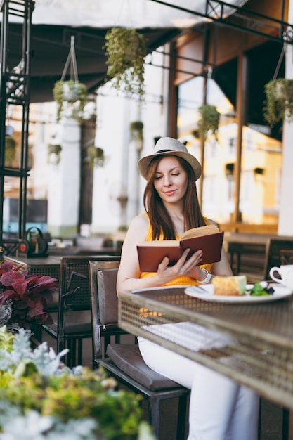 Frau im Straßencafé im Freien, die am Tisch mit Hut sitzt, ein Buch mit einer Tasse Cappuccino, Kuchen liest, sich in der Freizeit im Restaurant entspannt?