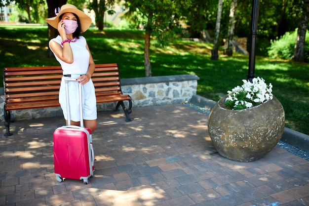 Frau im Strohhut und in der rosa Schutzmaske, im Park im Freien mit einem Koffer, auf einem Handy sprechend, Leben während der Coronavirus-Pandemie, Eröffnungsflugreise, Reisekonzept.