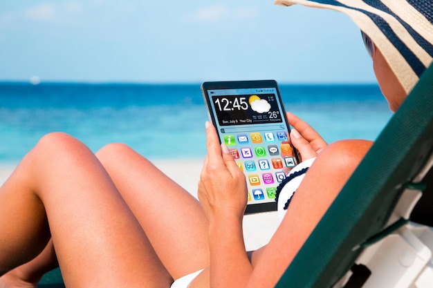 Frau im Urlaub liegt in einer Sonnenliege am Strand mit einem Tablet in den Händen.