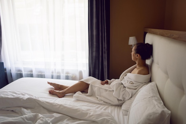 Frau im weißen Gewand sitzt nach dem Duschen auf einem Bett am Fenster in einer Hotelwohnung