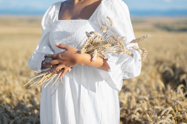 Frau im weißen Kleid steht im Feld mit Weizen Person in Händen halten Bündel reife Ährchen erntet...