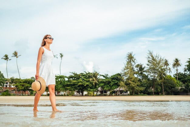 Frau im weißen Kleid und Strohhut geht bei Sonnenuntergang an einem tropischen Strand spazieren