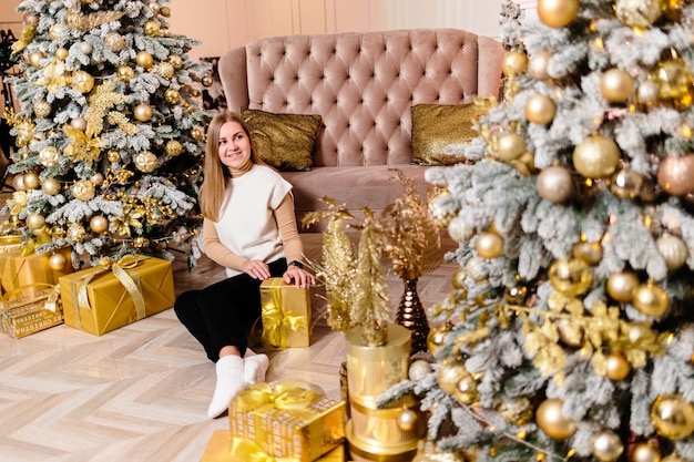 Frau im weißen Pullover, warmer und gemütlicher Abend in einem weihnachtlichen Interieur, Weihnachtsbaum mit Lichtern geschmückt, Girlande, die drinnen aufleuchtet. Weicher Fokus