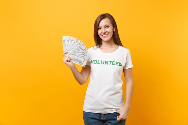 Frau im weißen T-Shirt mit schriftlicher Aufschrift grüner Titel Freiwilliger hält viele Dollar-Banknoten, Bargeld isoliert auf gelbem Hintergrund. Freiwillige kostenlose Hilfe, Konzept der Wohltätigkeitsarbeit.