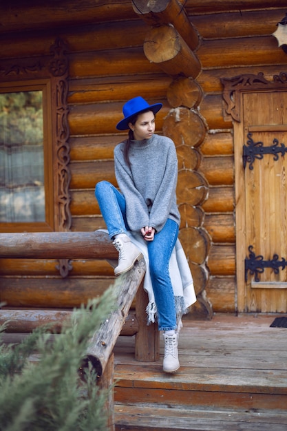 Frau in blauem Hut und Pullover steht im Herbst an der Tür des Holzhauses aus Baumstämmen