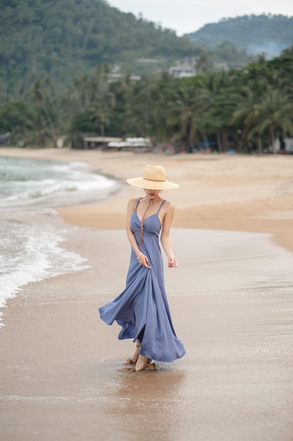 Frau in blauem Kleid und Strohhut geht am Strand spazieren.