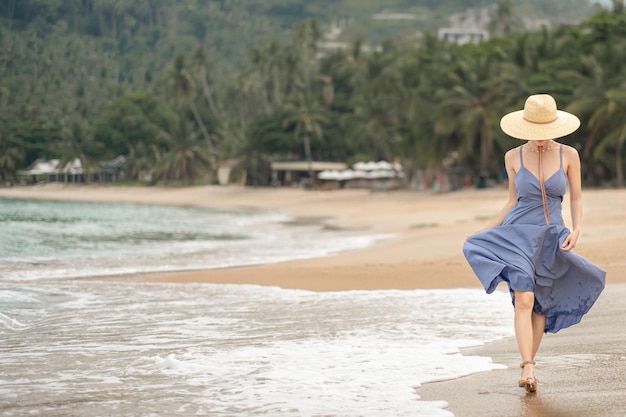 Frau in blauem Kleid und Strohhut geht am Strand spazieren.