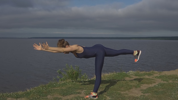 Frau in blauer Sportbekleidung, die auf einem Bein steht und die Arme nach vorne gestreckt, Yogapraxis im Freien in Aktion, Rückansicht, bewölkter Himmel und See im Hintergrund. Yogi, Gleichgewichtstraining, gesunder Lebensstil