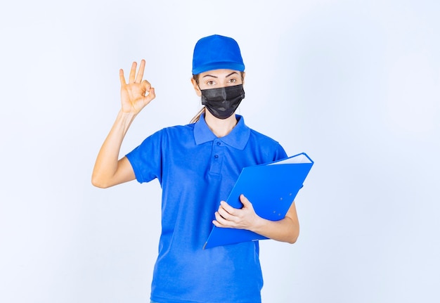 Frau in blauer Uniform und schwarzer Gesichtsmaske, die einen blauen Ordner hält und sich positiv und freundlich fühlt.