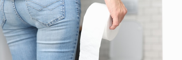 Frau in Blue Jeans steht mit dem Rücken und hält Toilettenpapierrolle in der Hand vor Toilettennahaufnahme. Urologiekonzept für häufiges Wasserlassen.