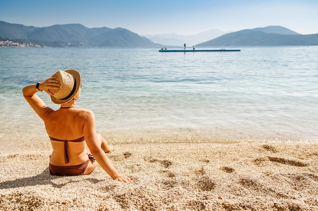 Frau in brauner Badebekleidung und Strohhut sitzt am Strand von Montenegro und genießt den sonnigen Tag