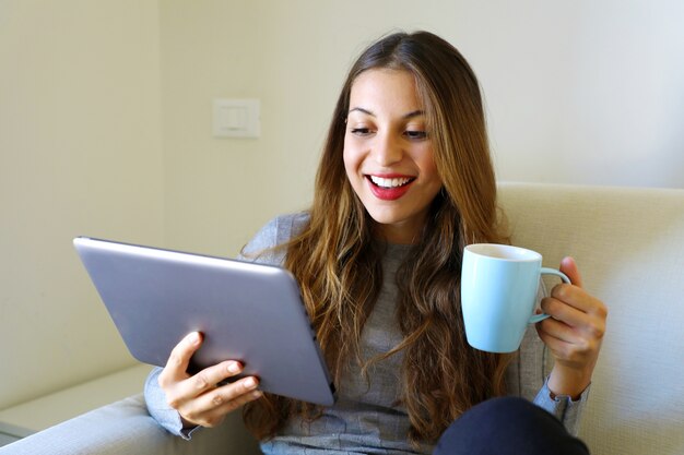 Frau in den gemütlichen Kleidern des Hauses, die auf einem Sofa unter Verwendung der Tablette sitzen und Tasse halten