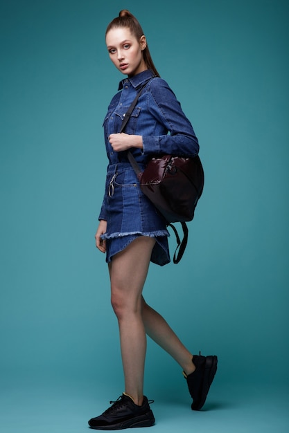 Frau in Denim Jeans Kleid schwarze TurnschuheRucksack auf türkisblauem Hintergrund Studio Shot