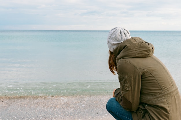 Frau in der Jacke und in Hut, die das Meer während des Winters betrachten, geht auf den Strand