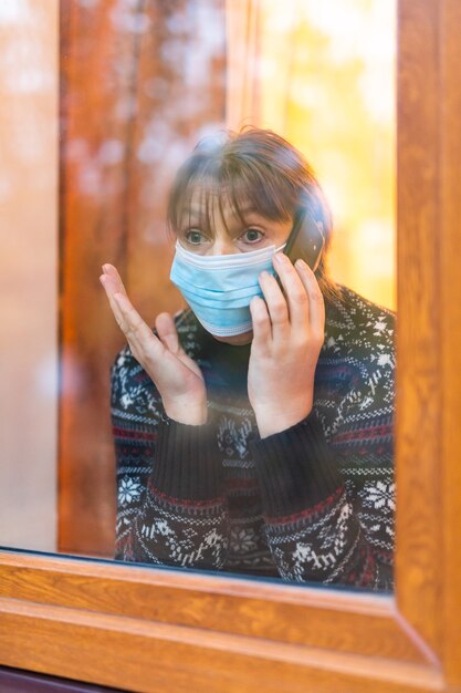 Frau in der medizinischen Maske, die durch Fenster mit Hoffnung schaut