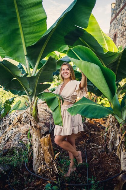 Frau in der Nähe von grünen Blättern von Bananenbüschen in der Natur in einem Park an einem tropischen Ort, sie trägt einen beigen Rock und eine Bluse