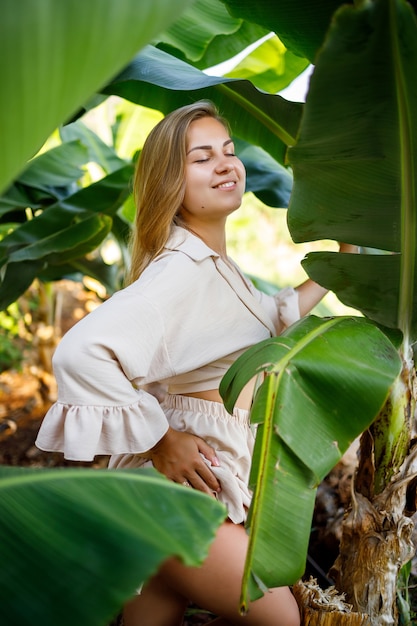 Frau in der Nähe von grünen Blättern von Bananensträuchern in der Natur in einem Park an einem tropischen Ort, sie trägt einen beigefarbenen Rock und eine Bluse