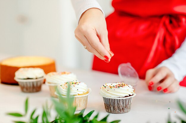 Frau in der roten schürze verziert mit farbigen streuseln cupcakes in der küche zu hause.