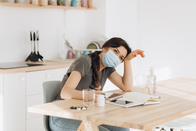 Frau in der Schutzmaske, die in der Faust hustet krank von Coronavirus-Virusinfektion, die Coronavirus verbreitet, das Mund und Nase bedeckt. Kopfschmerzkranker Patient, der in der Küche zu Hause Quarantäne sitzt