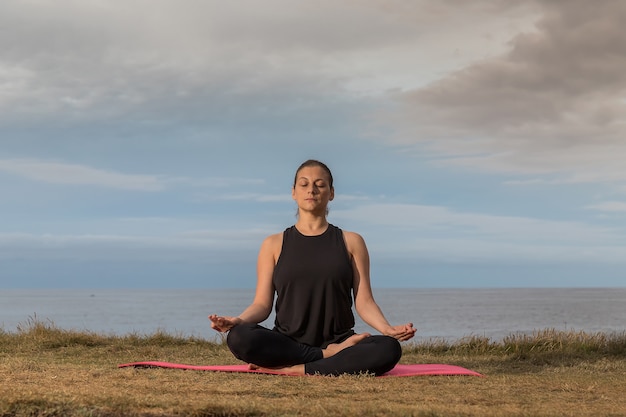 Frau in der Sportbekleidung, die Yoga draußen auf einer rosa Matte mit dem Meer tut