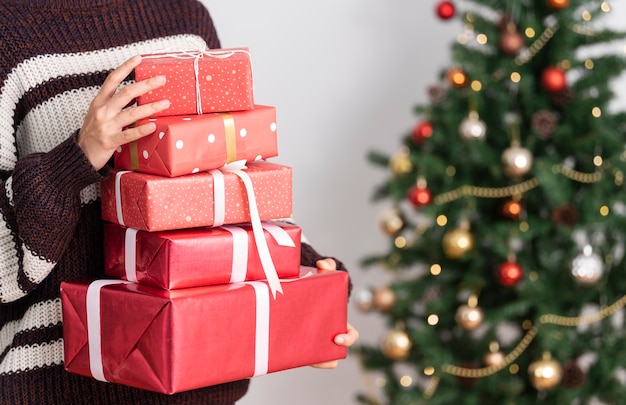 Frau in der Strickjacke, die Geschenkbox nahe Weihnachtsbaumhintergrund hält.