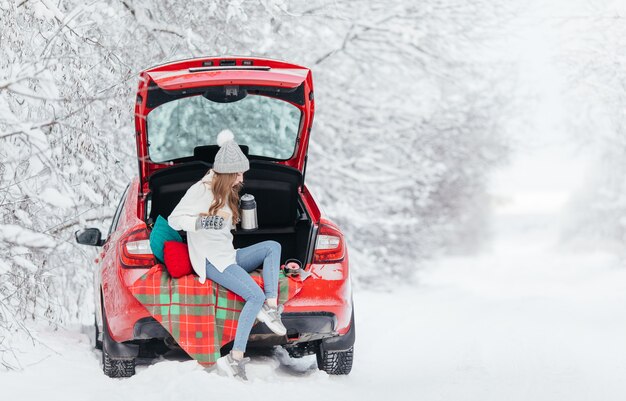 Frau in der warmen Kleidung, die im Winterwald sitzt, während sie sich auf das Auto stützt und Tasse Kaffee hält.