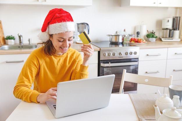 Frau in der Weihnachtsmütze, die online einkauft und mit Goldkreditkarte zahlt