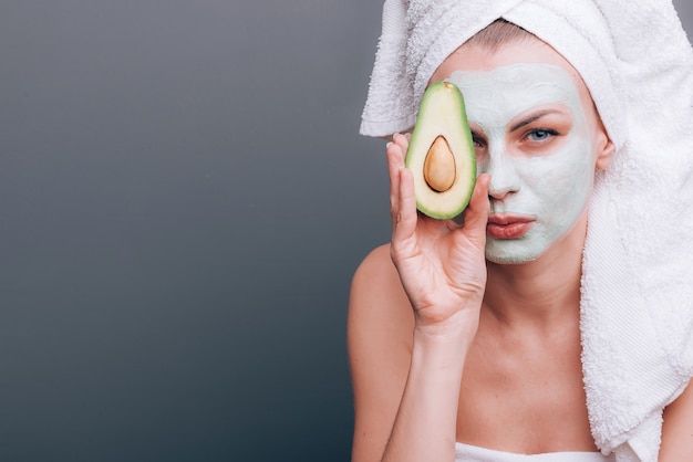 Frau in ein Handtuch gewickelt mit einer kosmetischen Maske im Gesicht und Avocado in den Händen
