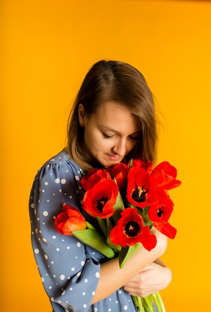 Frau in einem blauen Kleid hält rote Tulpen und schnüffelt Blumen an einer gelben Wand