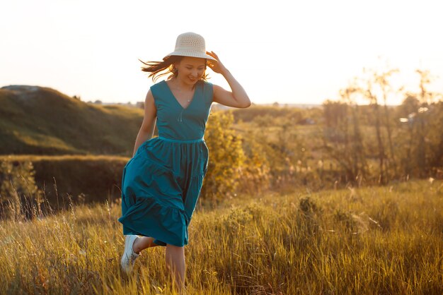 Frau in einem blauen Kleid und Panama läuft auf einer Sommerwiese bei Sonnenuntergang.