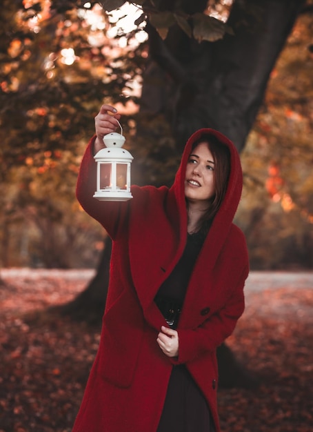 Frau in einem burgunderfarbenen Mantel mit Laterne im Herbstwald im viktorianischen Stil