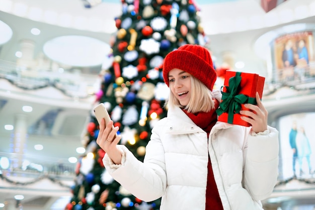 Frau in einem Einkaufszentrum hält ein Geschenk mit einer grünen Schleife in der Hand und macht lachend ein Selfie